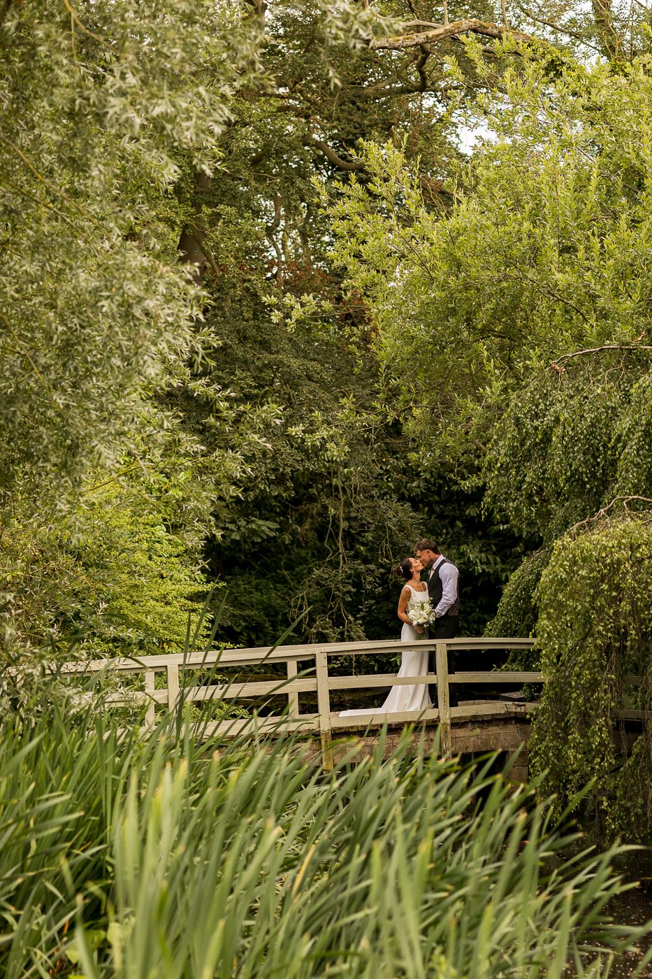 A bride and groom share a romantic kiss on a wooden bridge surrounded by lush green trees in a garden setting.