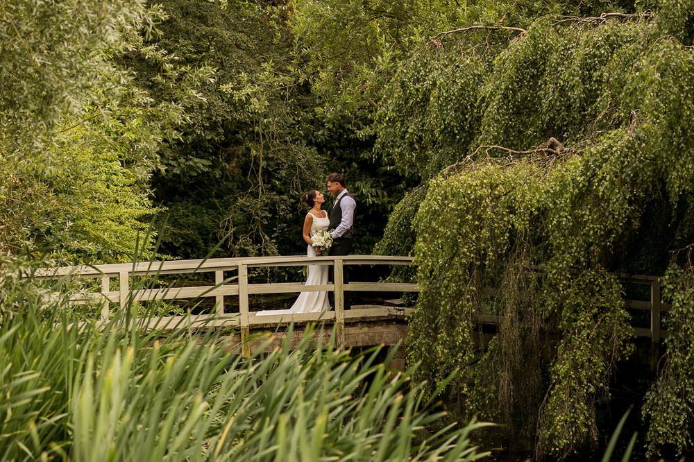 A bride and groom embrace on a wooden bridge surrounded by lush green weeping willow trees in a garden setting.