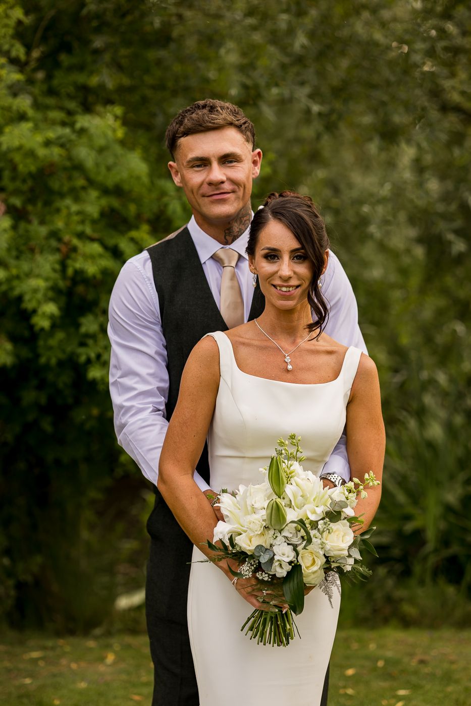 Smiling bride holding white floral bouquet stands with groom in vest and tie outdoors at wedding ceremony.