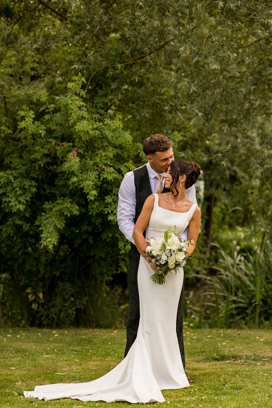 Bride and groom embrace in lush garden setting, bride in elegant white gown holding bouquet of white and green flowers.