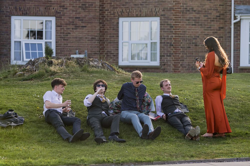 Four young men in formal wear sit on grass while a woman in an orange gown stands talking to them outdoors.
