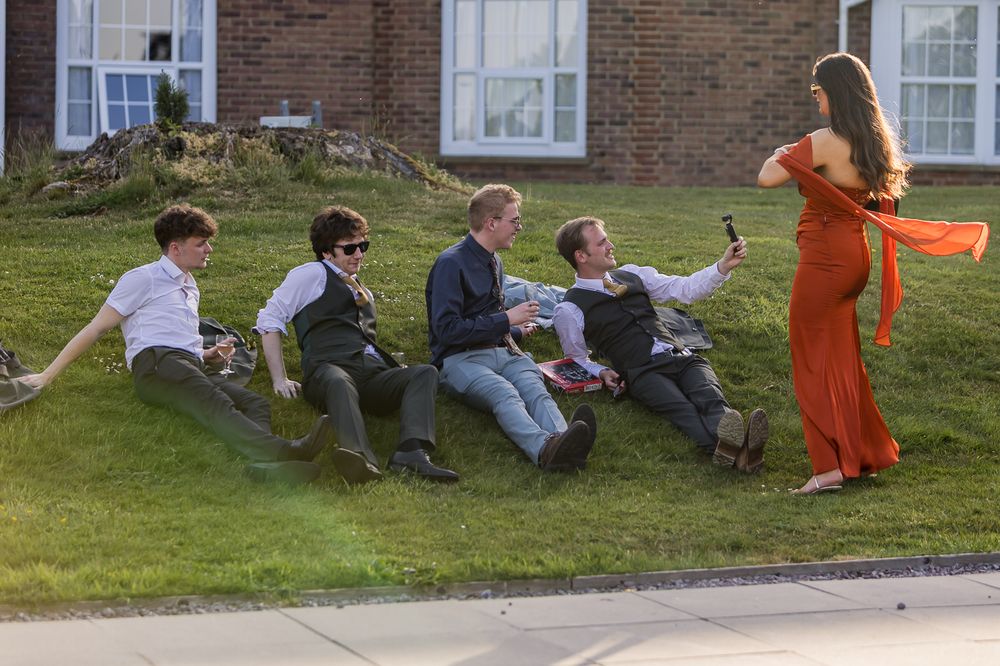 Four men lounging on a grassy slope take a selfie with a woman in an elegant orange evening gown outside.