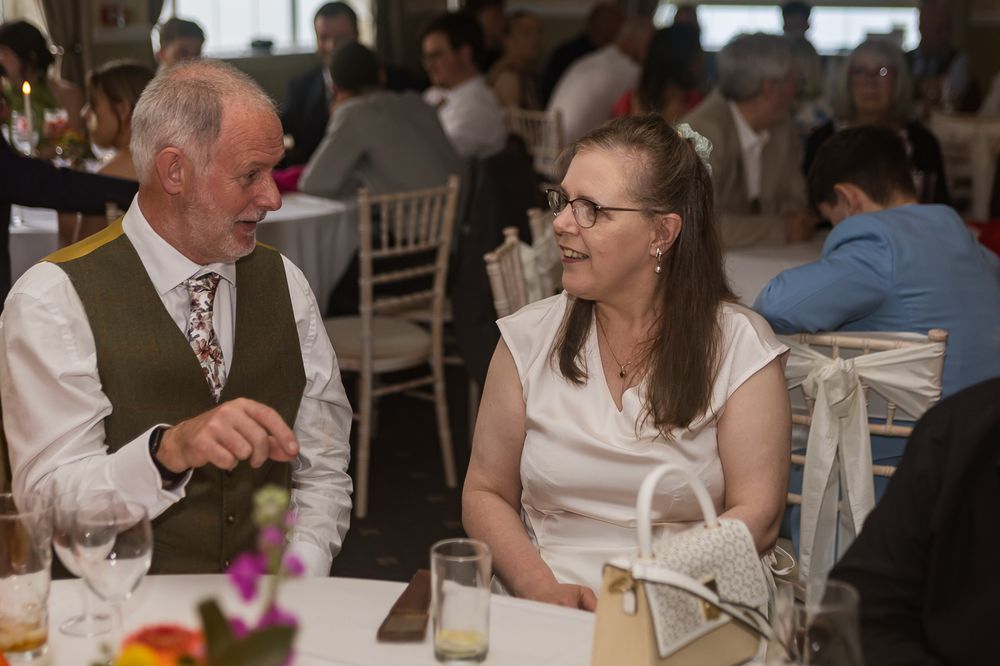 Elderly man in vest and woman in white dress engaged in conversation at a formal wedding reception dinner.