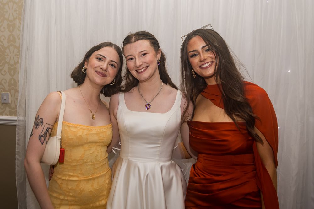 Three women pose together at a wedding, one wearing a white bridal gown flanked by guests.