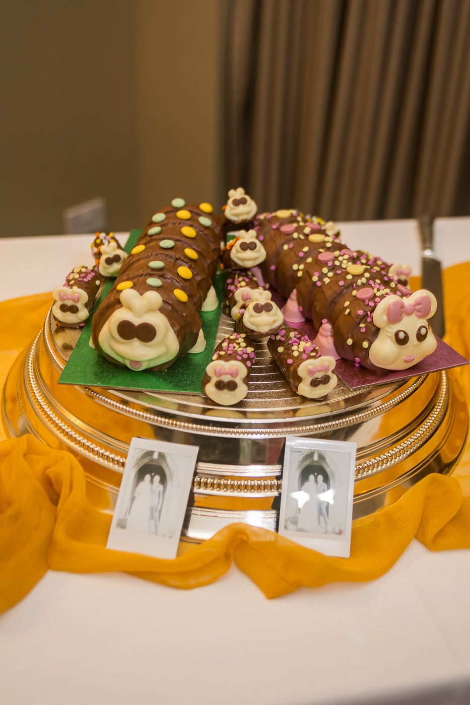 Two caterpillar-shaped chocolate celebration cakes on a silver platter with yellow fabric, decorated with colorful candy buttons.