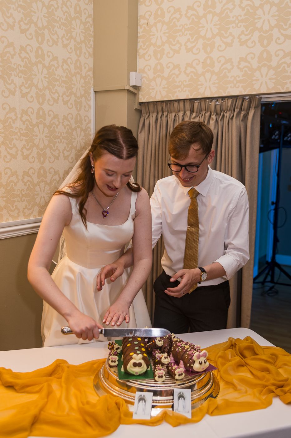 Bride and groom cutting a decorative wedding cake on a gold cloth-covered table in an elegant reception venue.