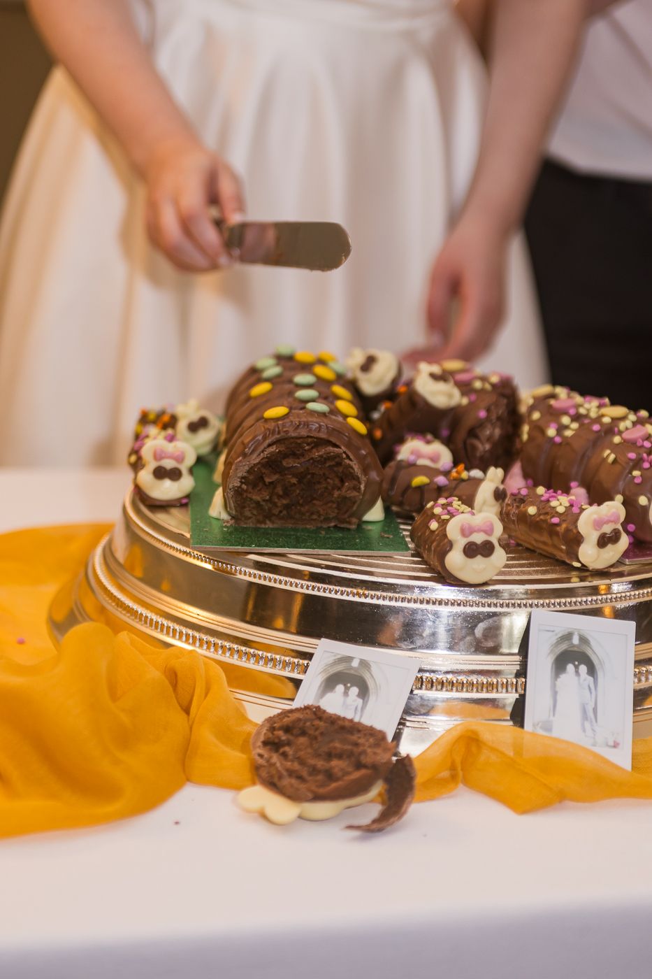 Chocolate yule log cake with colorful decorations on silver platter at wedding reception with couple cutting in background.
