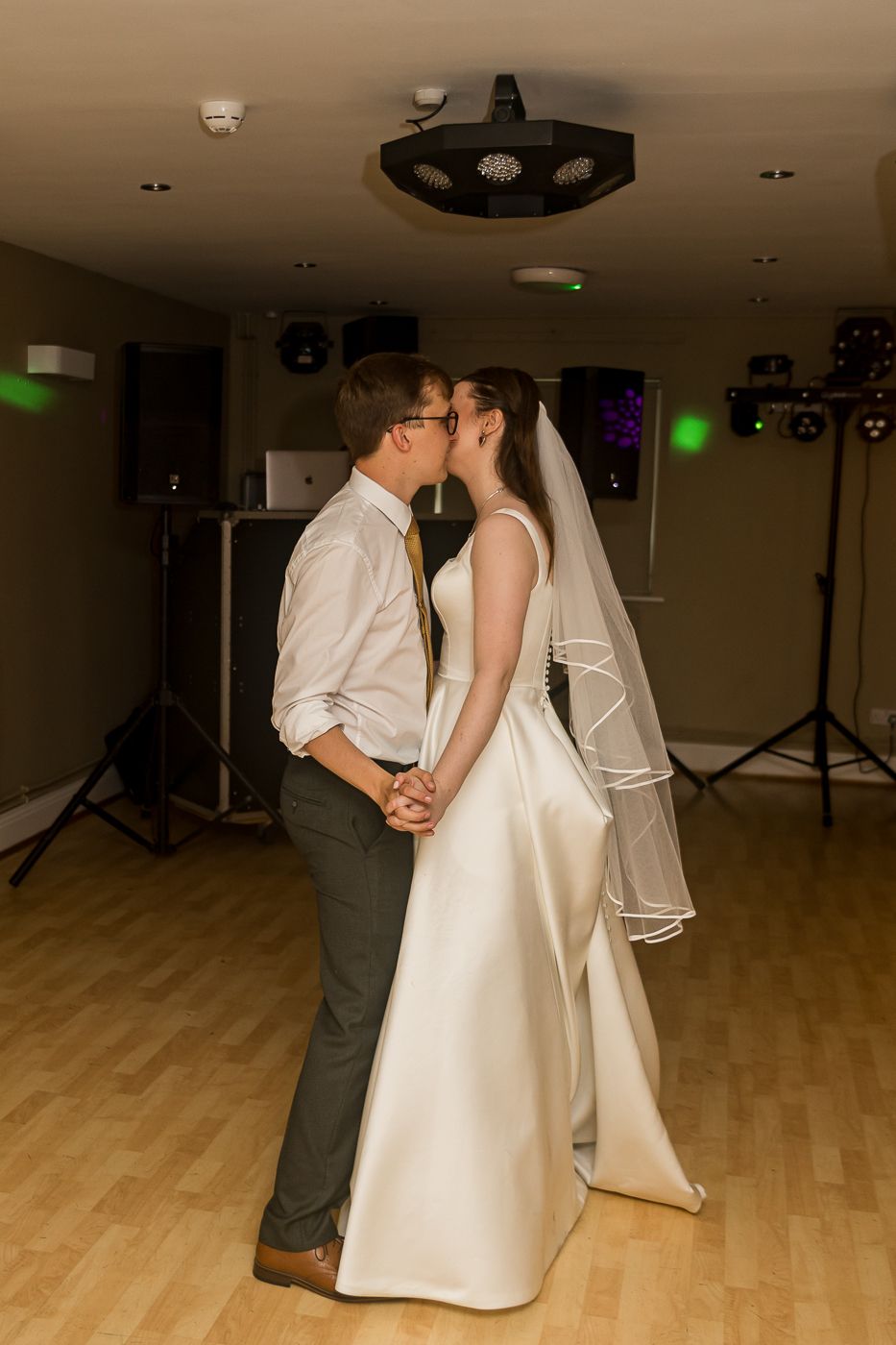 Bride and groom sharing their first dance kiss in a wedding reception venue with DJ equipment and colorful lights.