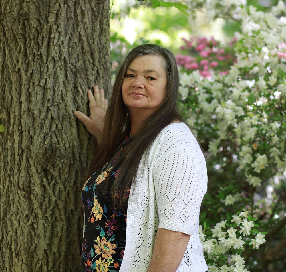 A woman with long dark hair leans against a tree surrounded by blooming white and pink flowers outdoors.