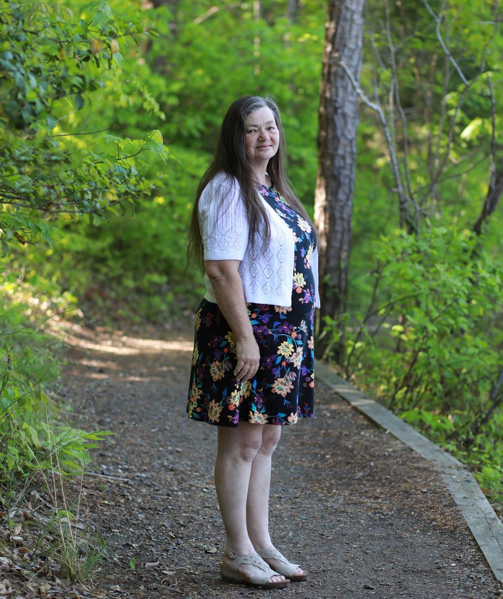 Woman in floral dress and white cardigan standing on a wooded path surrounded by lush green trees.