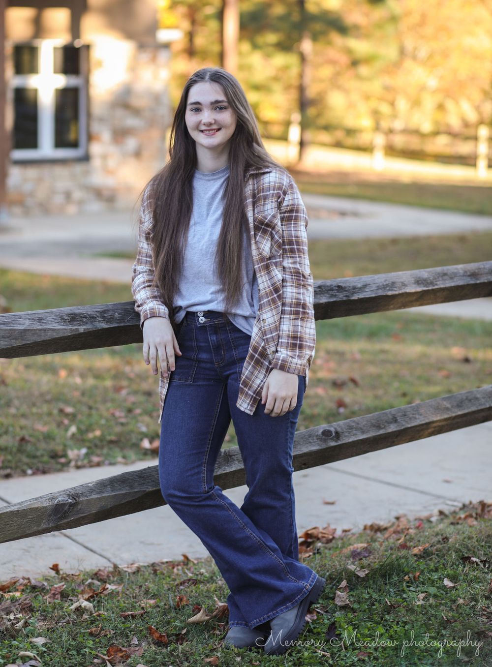 Young woman in plaid shirt and flared jeans posing by a wooden fence outdoors in autumn.