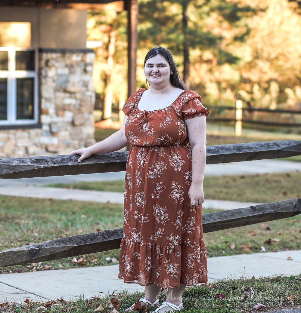Young woman in rust floral midi dress posing by wooden fence outdoors near stone building in autumn.