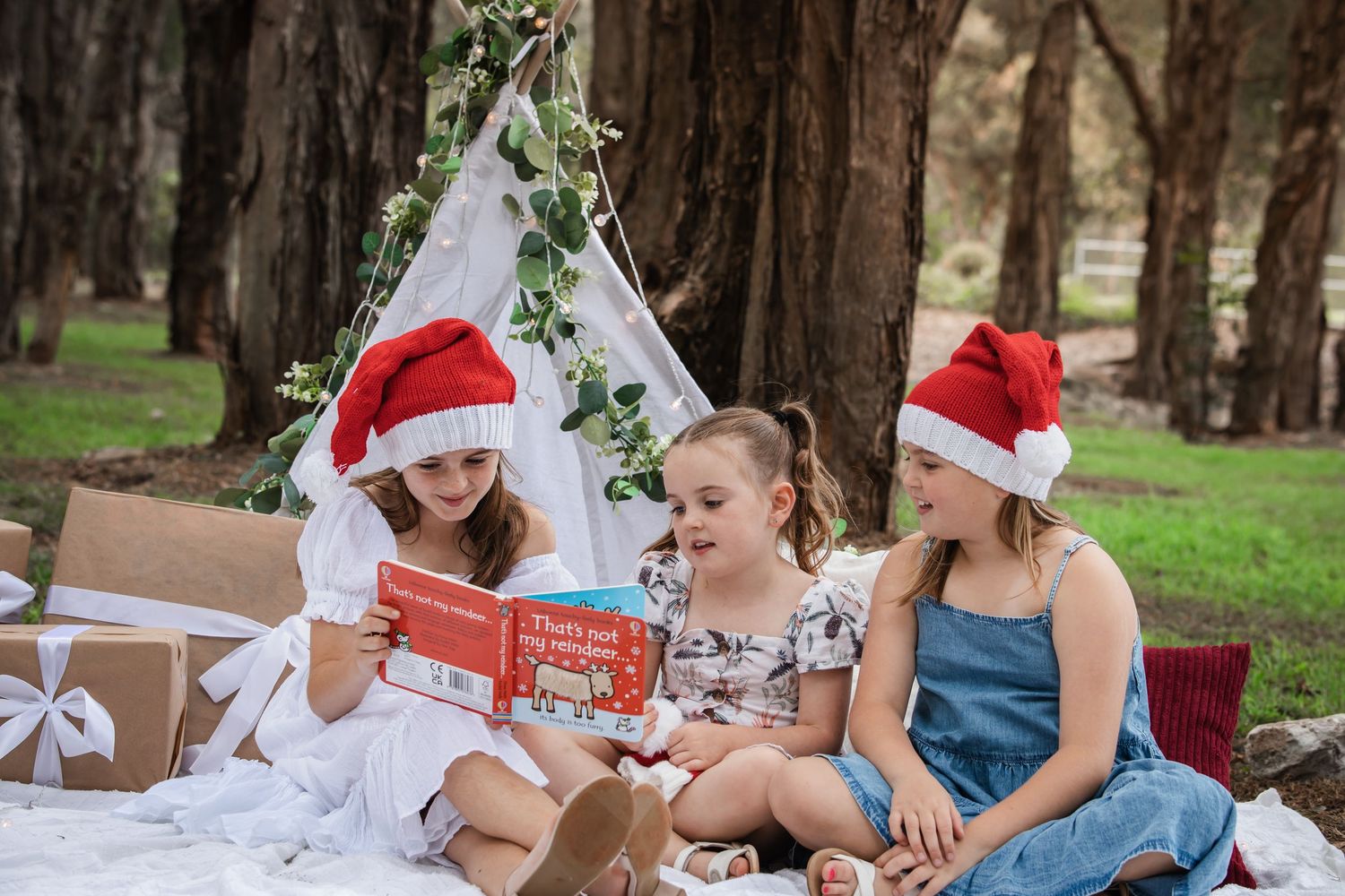 Three young girls, two in Santa hats, sit together on a picnic blanket reading a red Christmas book near a teepee tent and gift boxes.