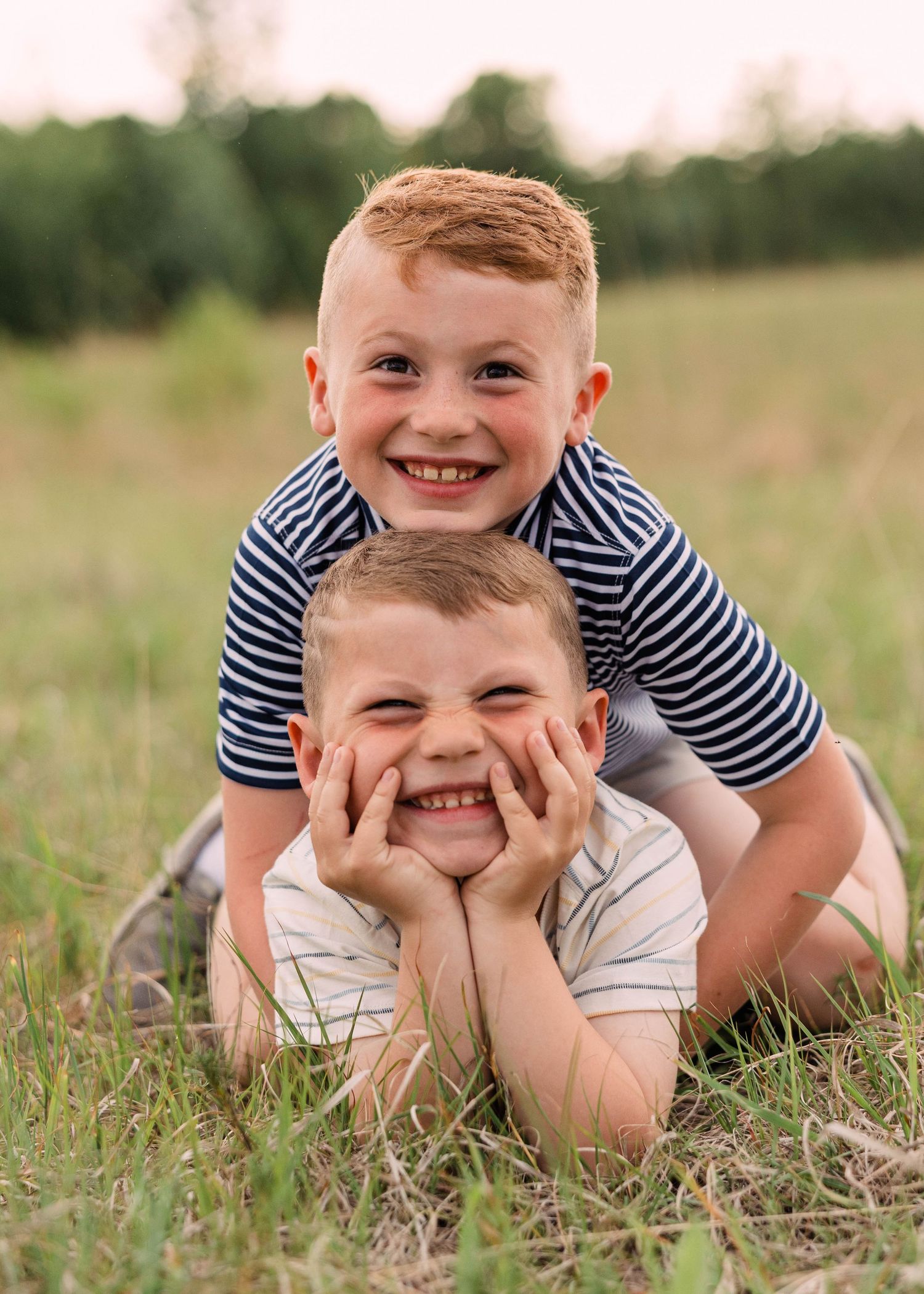 Two smiling young brothers in striped shirts pose playfully on a grassy field, one resting his chin in his hands.
