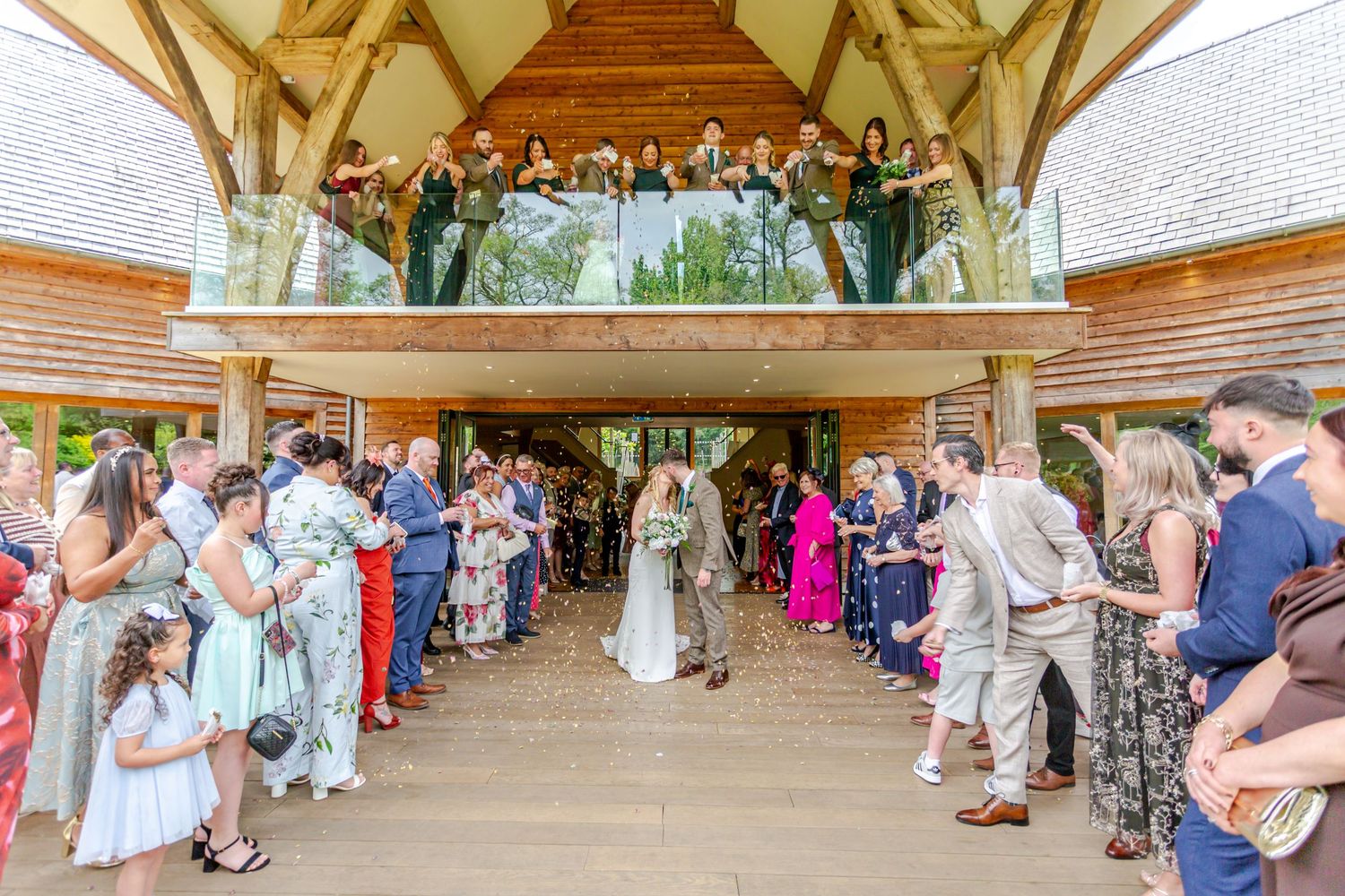 Bride and groom walk through cheering wedding guests in a stunning wooden venue with balcony onlookers.