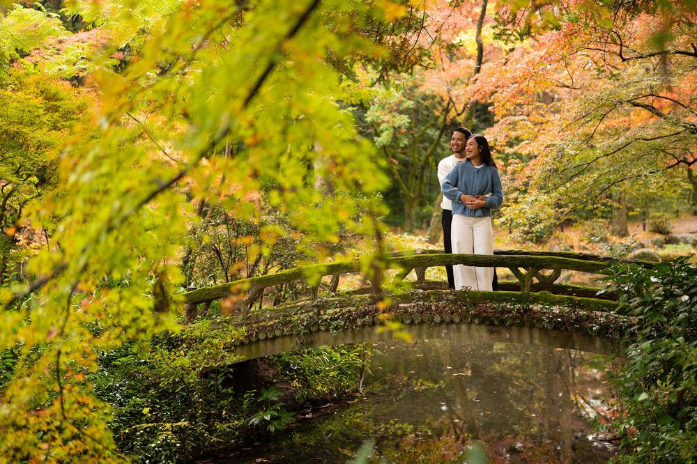 Couple embracing on a rustic wooden footbridge surrounded by colorful autumn maple foliage in a serene Japanese-style garden.