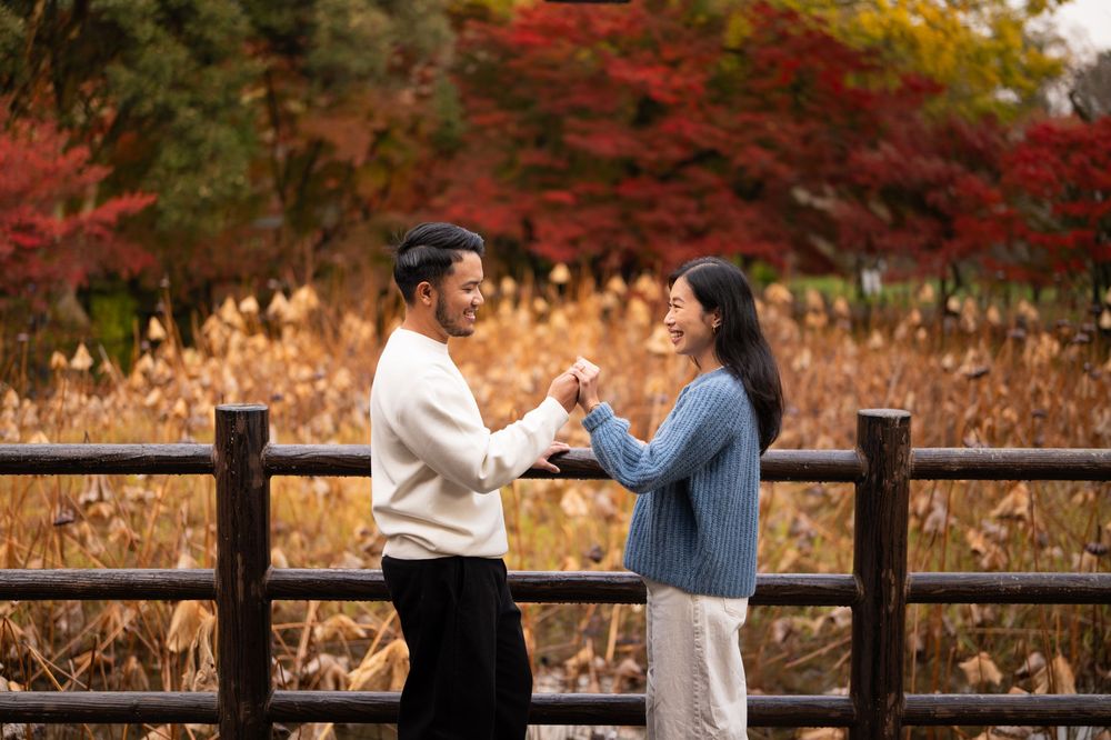 Smiling couple holding hands and facing each other beside a wooden fence, with golden grass and red autumn foliage in the background.