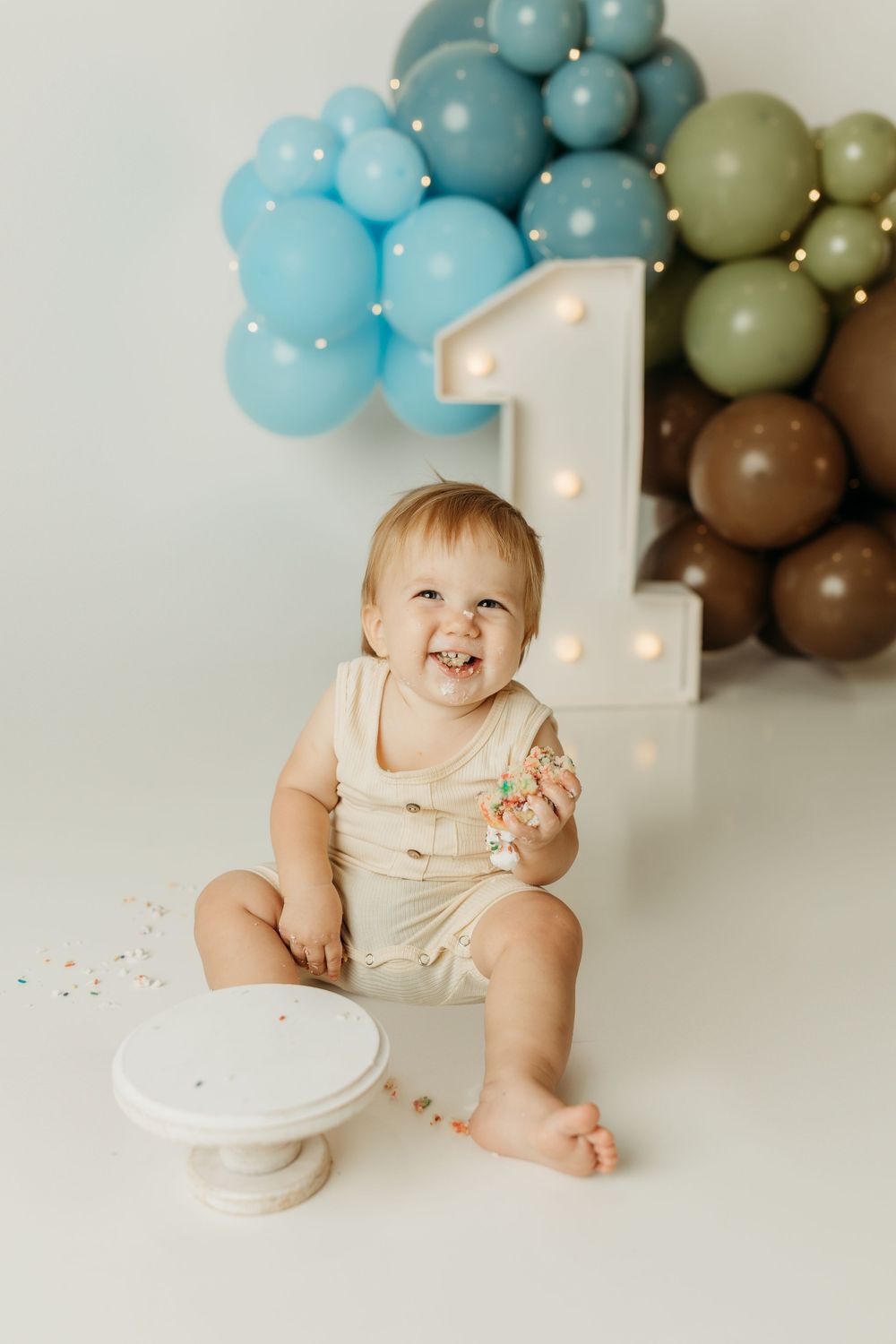 A happy baby celebrates their first birthday with cake, balloons, and a lit number one decoration in blue and green.