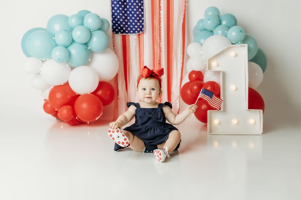 Baby girl in navy romper sits with American flag amid red, white, and blue balloons for 1st birthday.