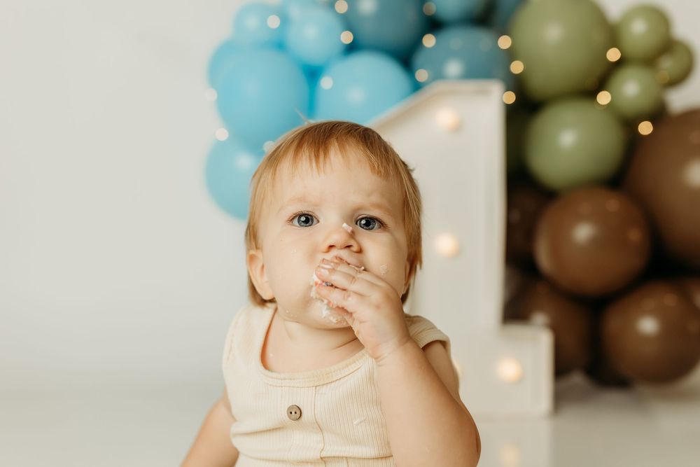 Baby celebrating first birthday with cake smash, surrounded by blue, green, and brown balloons with number one decoration.