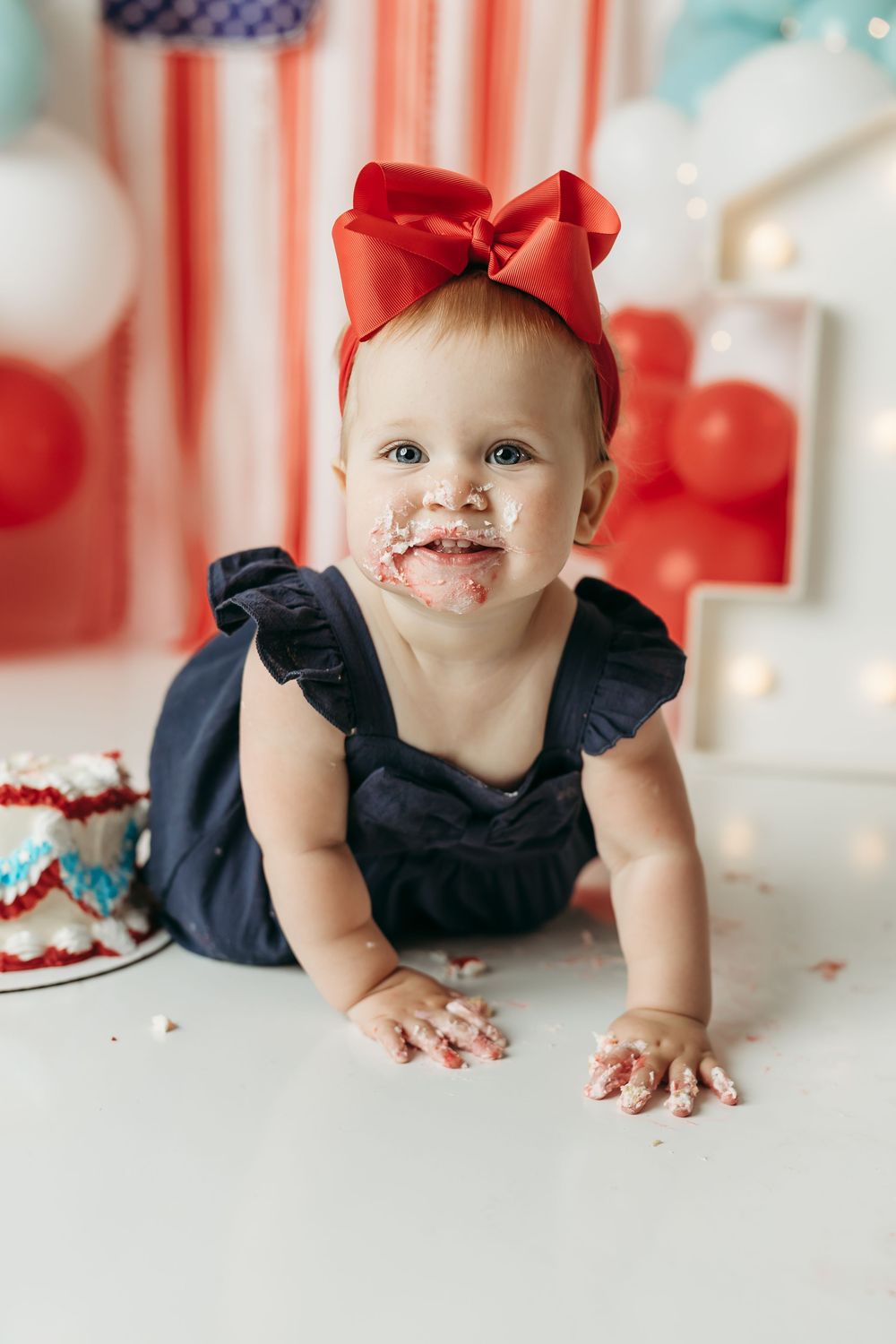 Baby girl in navy romper with red bow smashes first birthday cake, face covered in frosting, patriotic decor background.