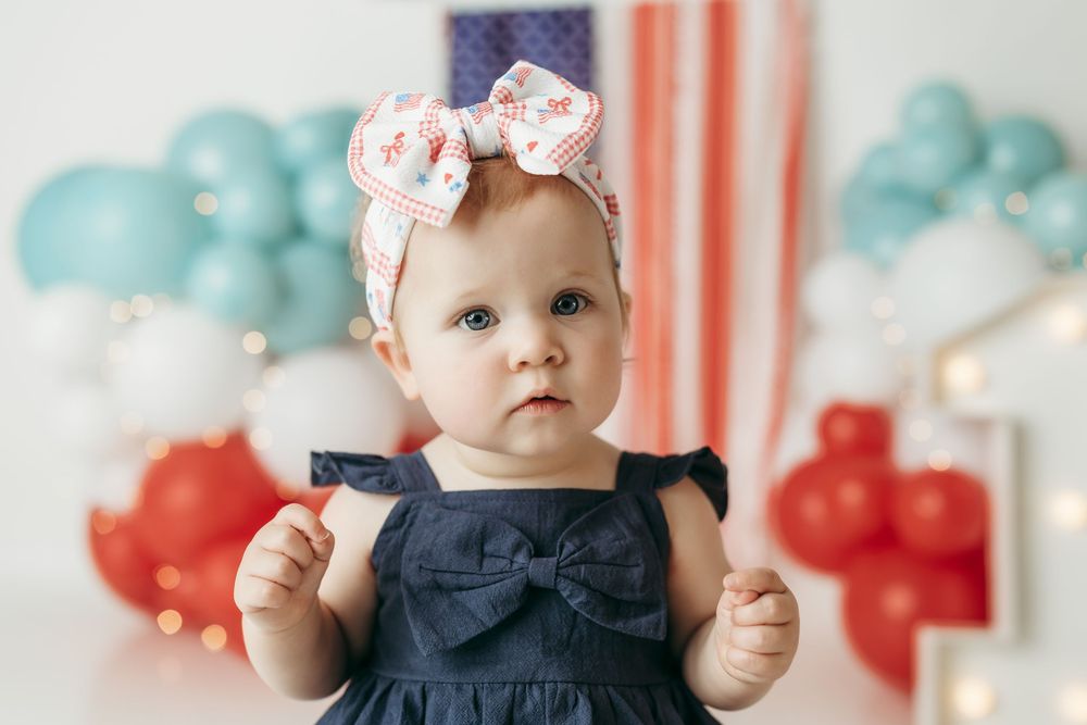 Adorable baby girl in navy bow dress and floral headband poses before American flag with red, white, and blue balloons.