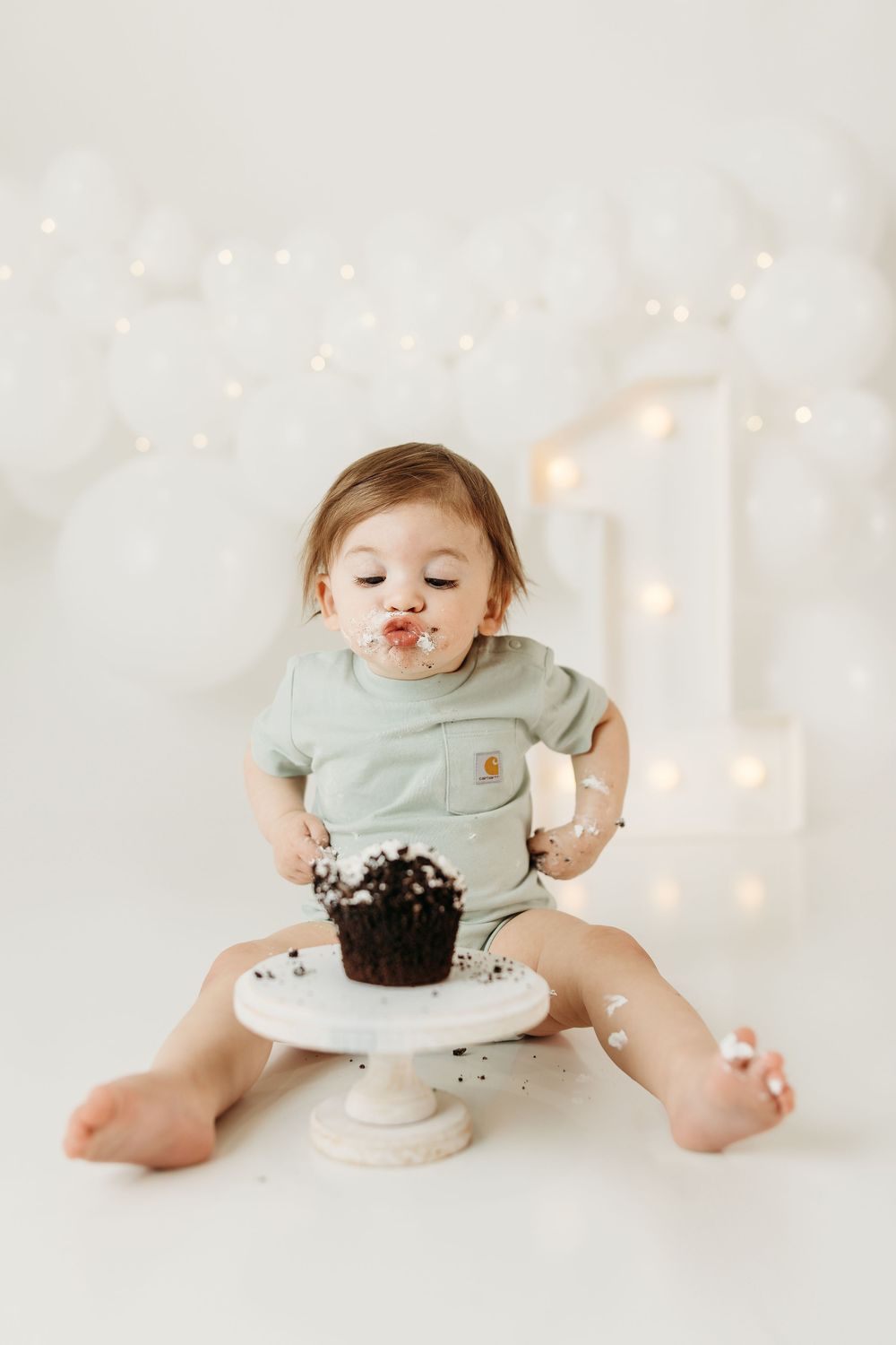Baby's first birthday cake smash with chocolate cupcake, sage green onesie, white balloons and fairy lights backdrop.