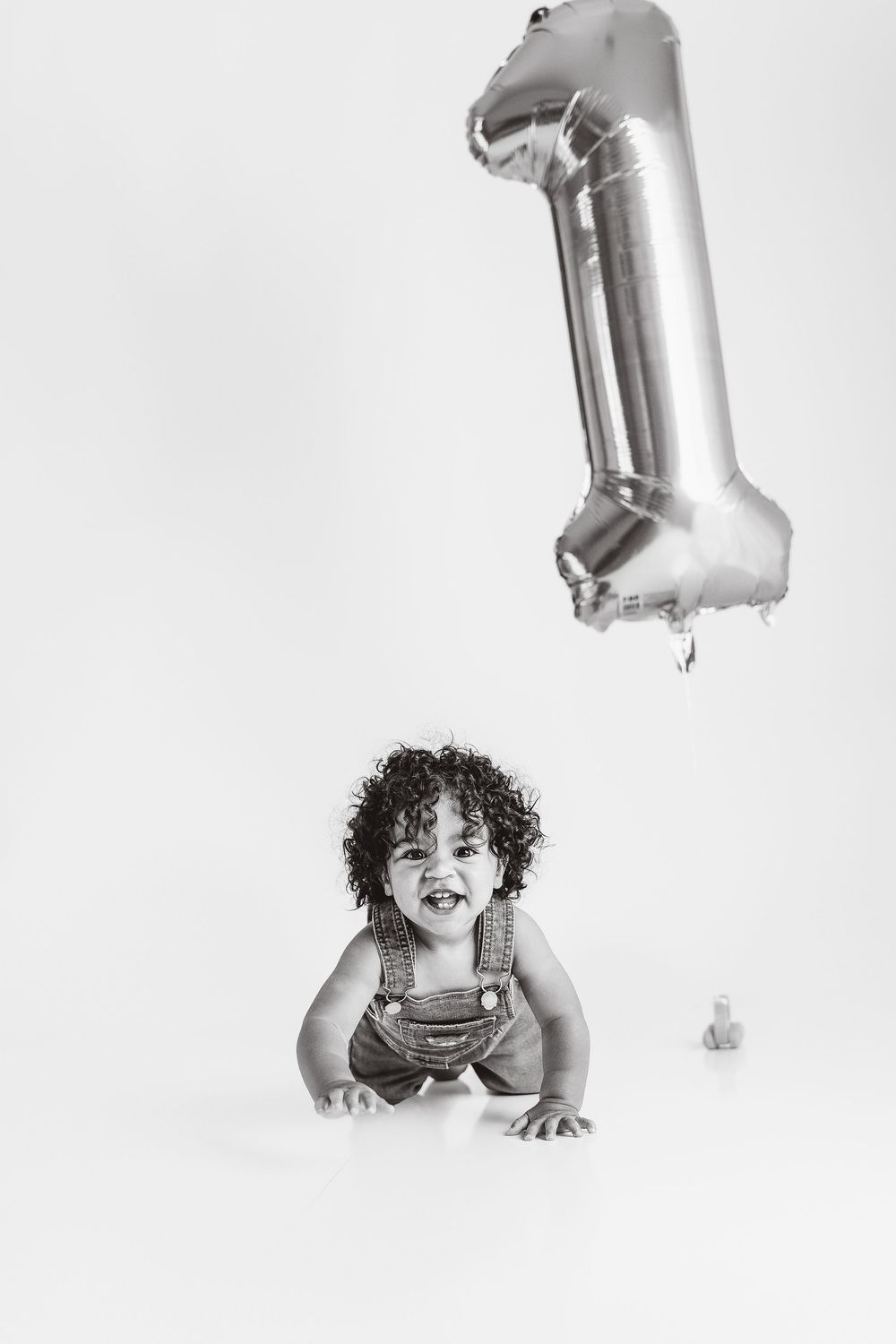Black and white photo of a happy baby crawling near a silver number 1 balloon, celebrating their first birthday.