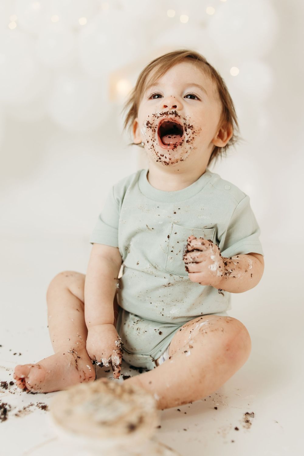 Messy baby in mint onesie enjoys first birthday cake smash, covered in chocolate frosting, sitting on white floor.
