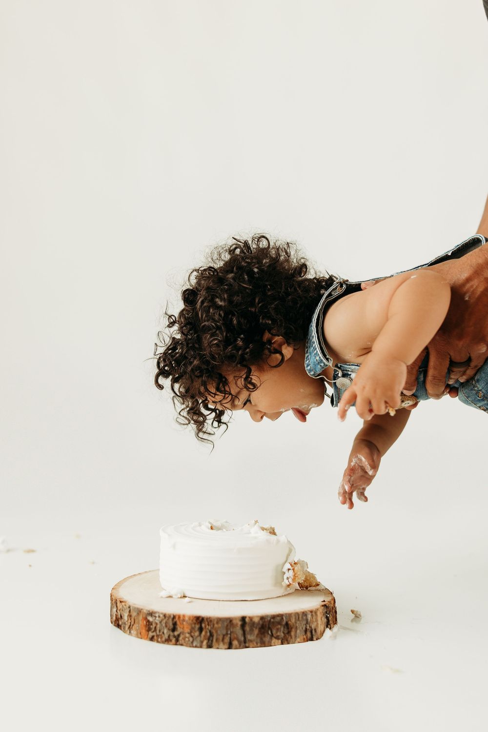 Curly-haired toddler leaning over a white smash cake on a rustic wood slice during a first birthday photoshoot.