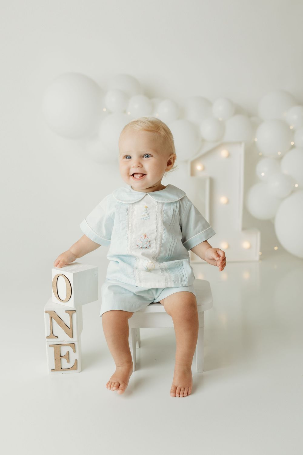 A smiling baby in a light blue outfit sits beside wooden ONE blocks and a lit number 1, surrounded by white balloons.