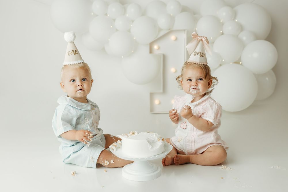 Two babies in party hats celebrate their first birthday with a white cake and balloon decorations.