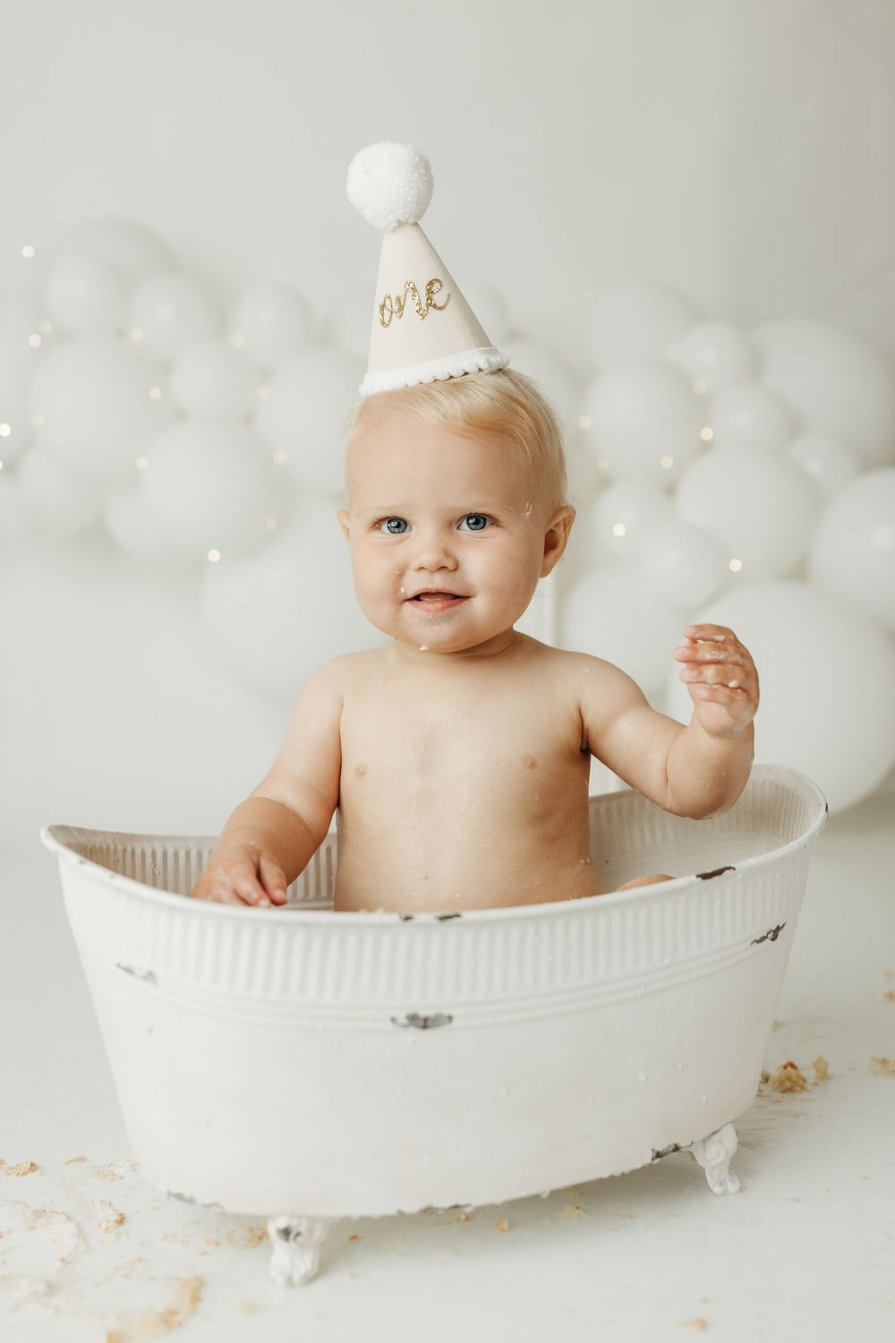 Baby in white tub wearing a birthday hat that says one, surrounded by white balloons for a first birthday cake smash.