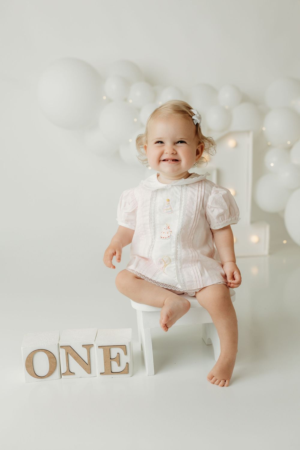 Smiling baby girl in pink outfit sitting beside ONE blocks at first birthday photo shoot with white balloons.
