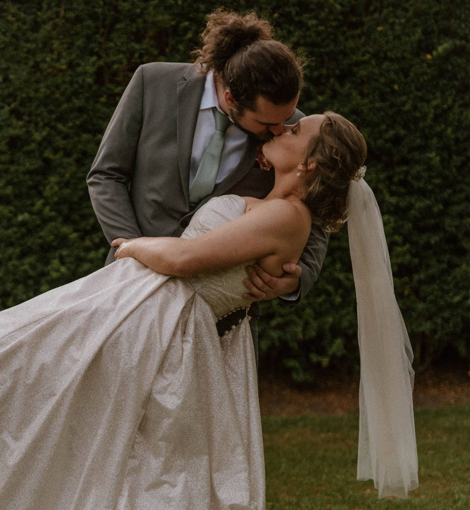 A groom in a gray suit dips his bride in a white gown and veil for a romantic kiss outdoors.