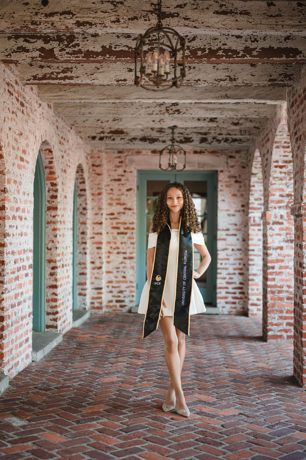 Graduate in a white dress and black graduation sash stands in a rustic brick archway corridor with hanging lanterns.