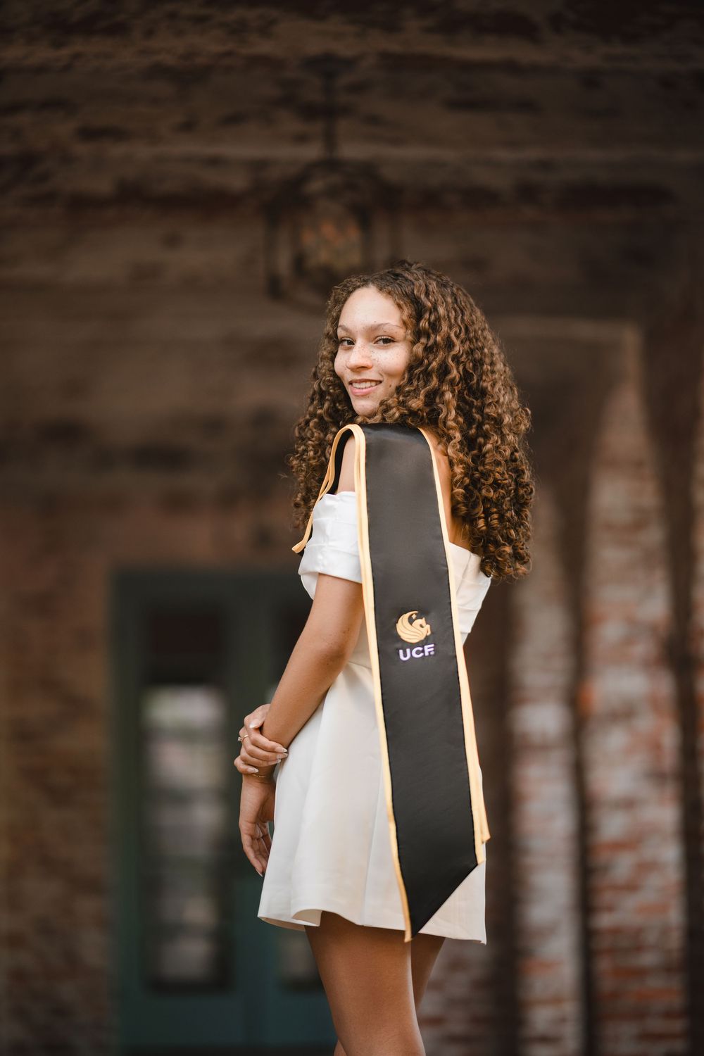 Smiling graduate with long curly hair wears a white dress and black UCR graduation stole while posing under a brick archway.