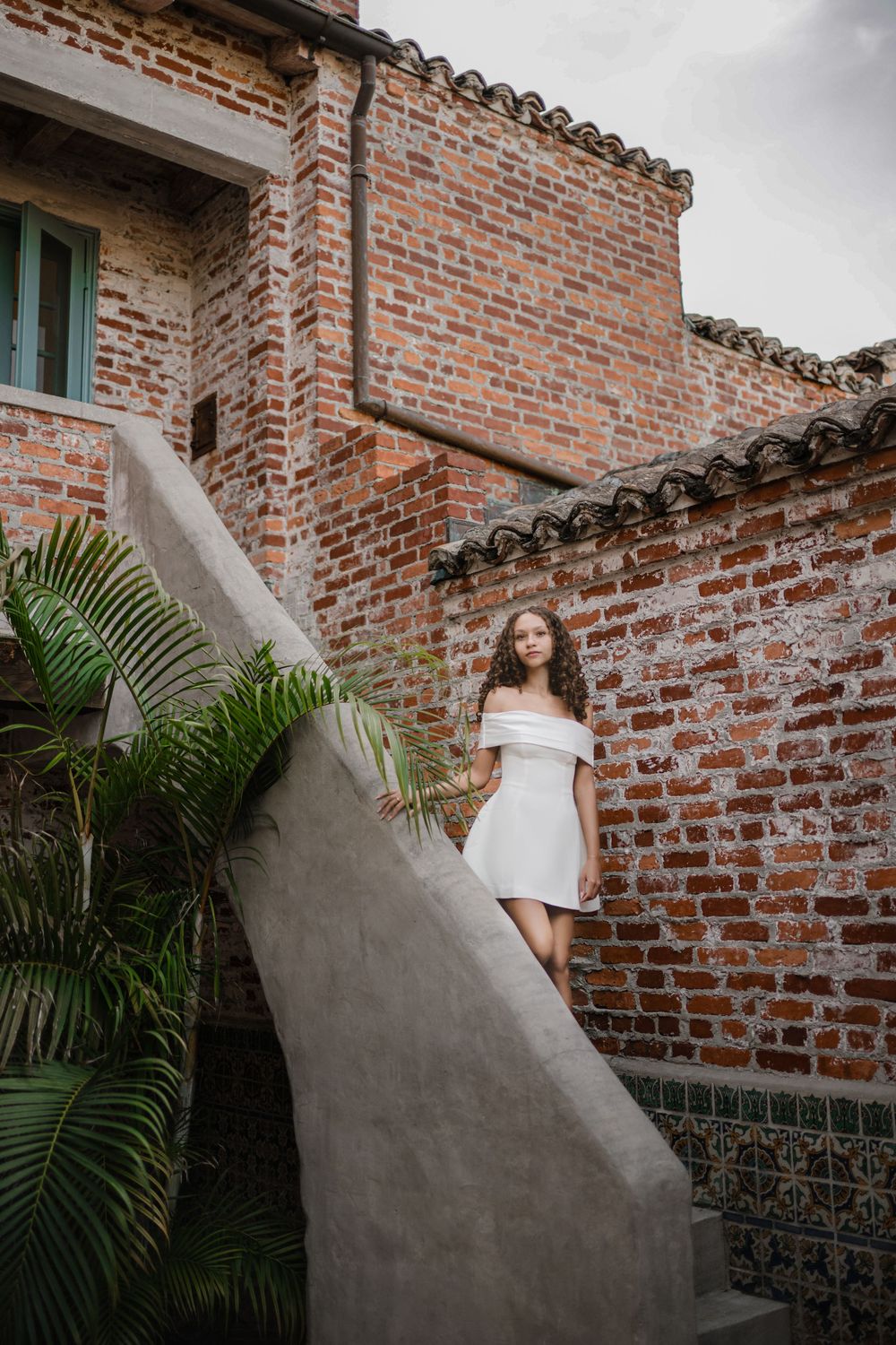 Young woman in a white off-the-shoulder dress standing on a concrete staircase beside a rustic red brick building with palm leaves.