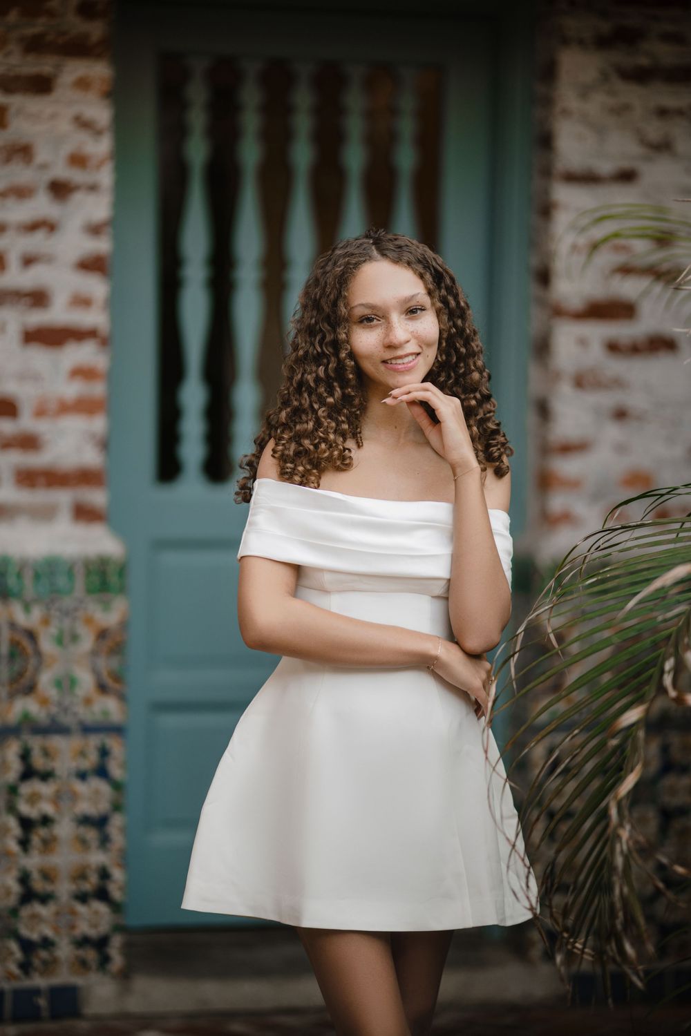 Smiling young woman with long curly hair poses in a white off-the-shoulder dress beside a teal door and brick wall with decorative tile.