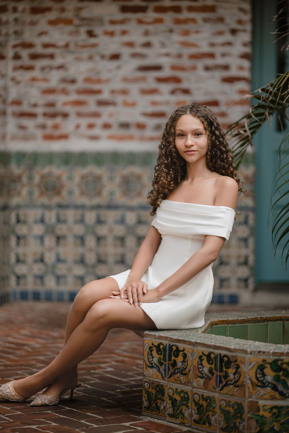 Young woman with long curly hair in a white off-the-shoulder dress sits on a decorative tiled ledge near a brick wall and palm leaves.
