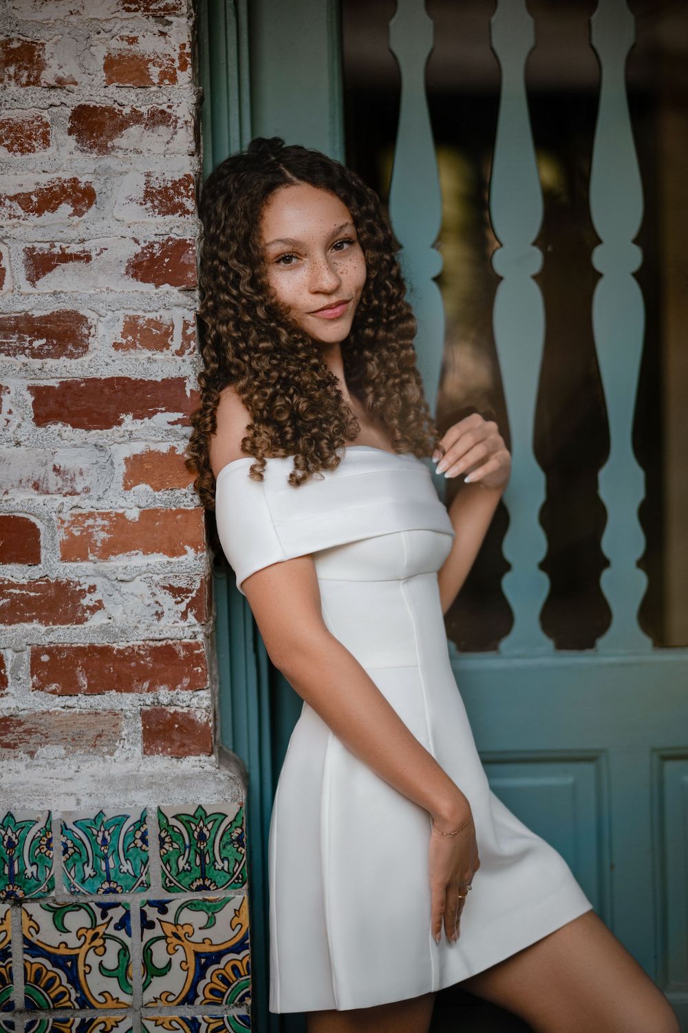 Young woman with long curly hair in a white off-the-shoulder dress posing beside a red brick wall and teal door with colorful mosaic tiles.