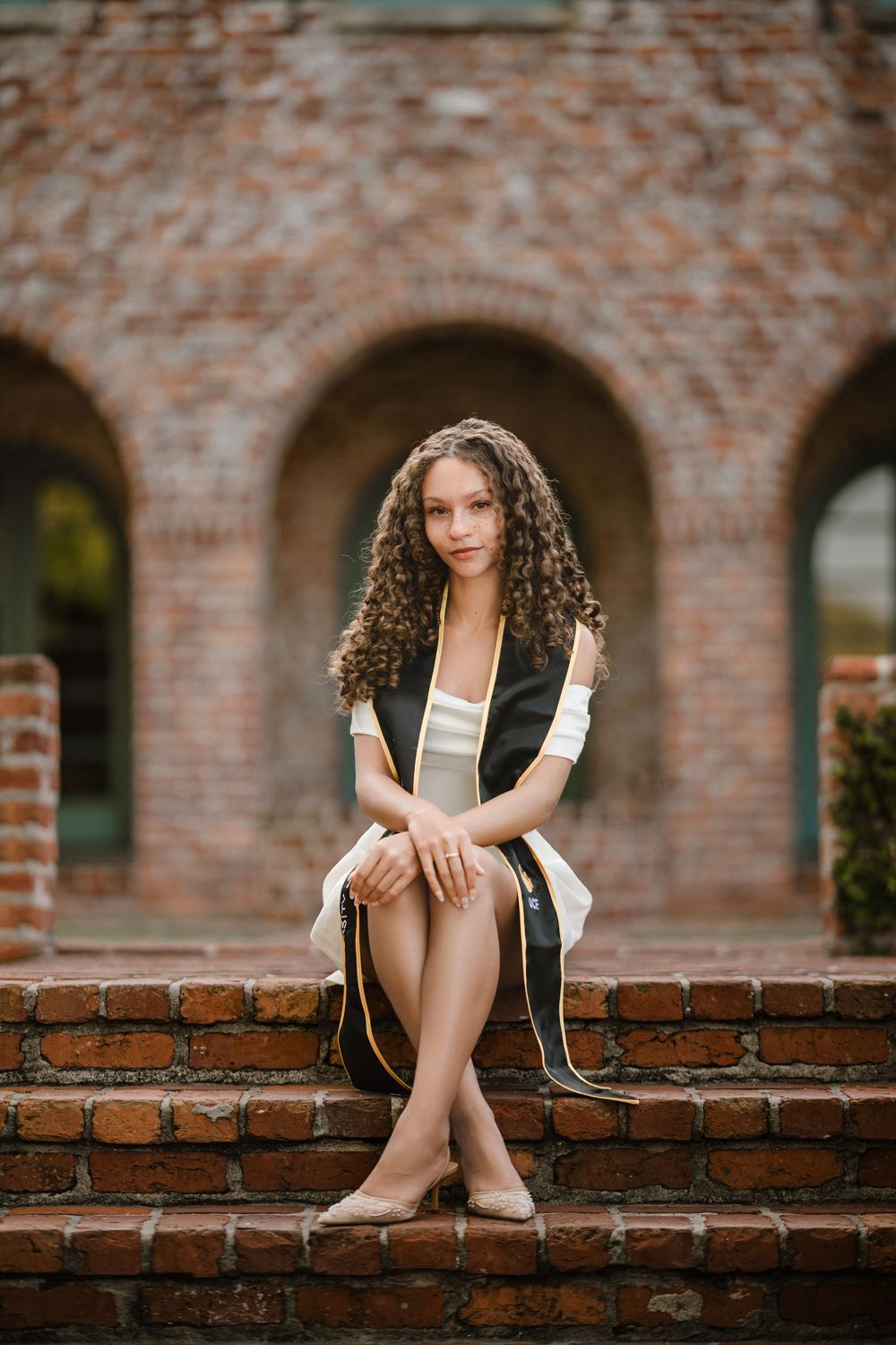 Young woman with long curly hair in a graduation stole sits on red brick steps in front of arched brick buildings for a grad portrait.