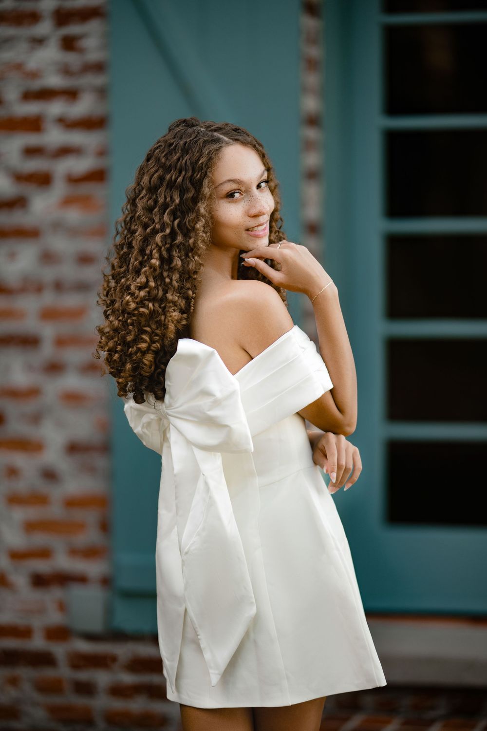 Young woman with long curly hair smiles over her shoulder in a white off-the-shoulder bow dress beside a teal door and brick wall.