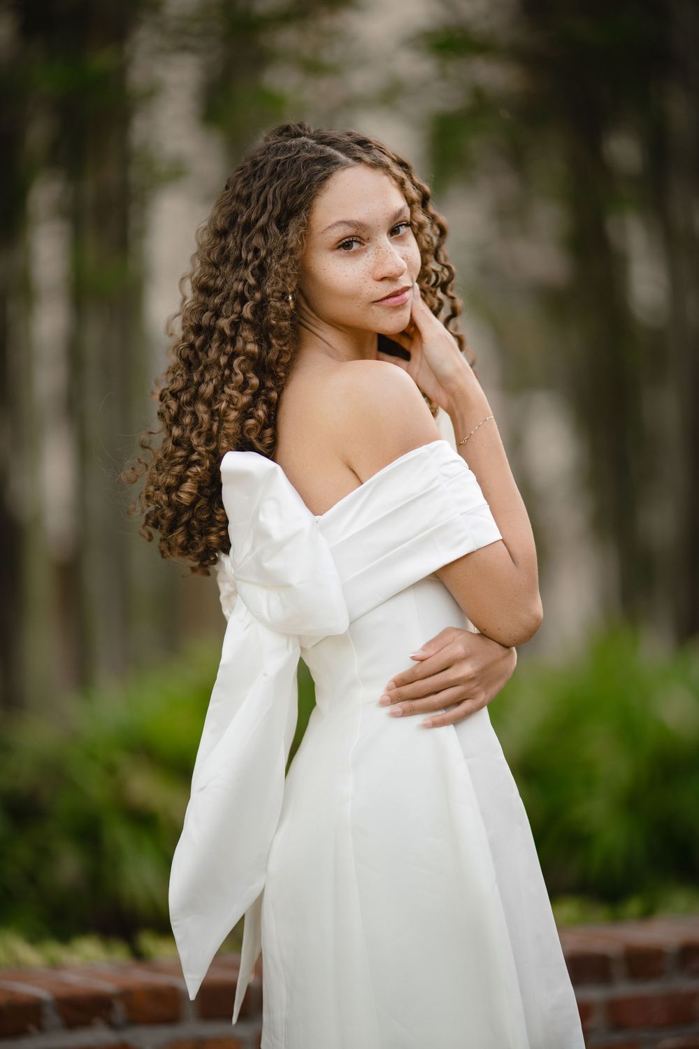 Young woman with long curly hair poses in a white off-the-shoulder dress with a large bow, standing near a brick wall and green foliage.
