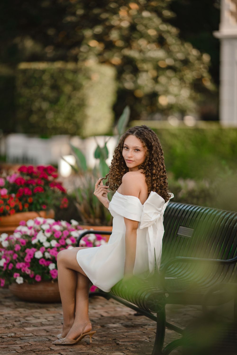 Young woman with long curly hair in a white off-the-shoulder dress sits on a black park bench beside pink and red flowers in a garden.