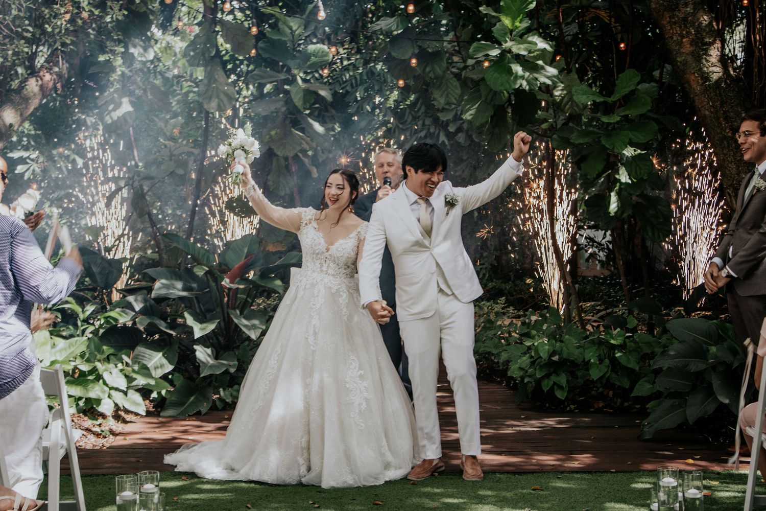 Bride in a short white mini dress and heels holds hands with a groom in a black suit on a brick path in front of a white building.