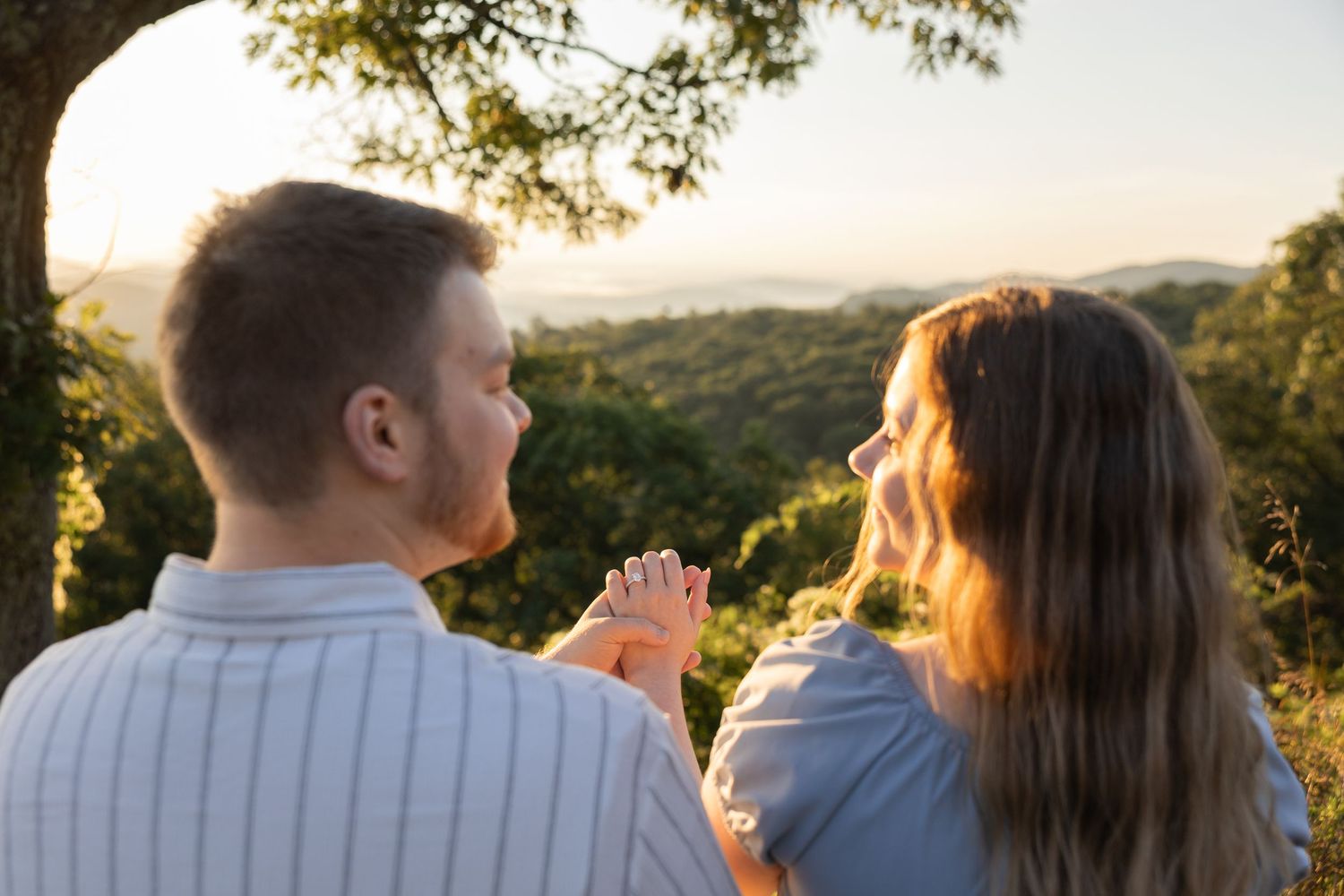 Couple holding hands at sunset overlooking lush green hills, romantic outdoor engagement photo in golden hour light.