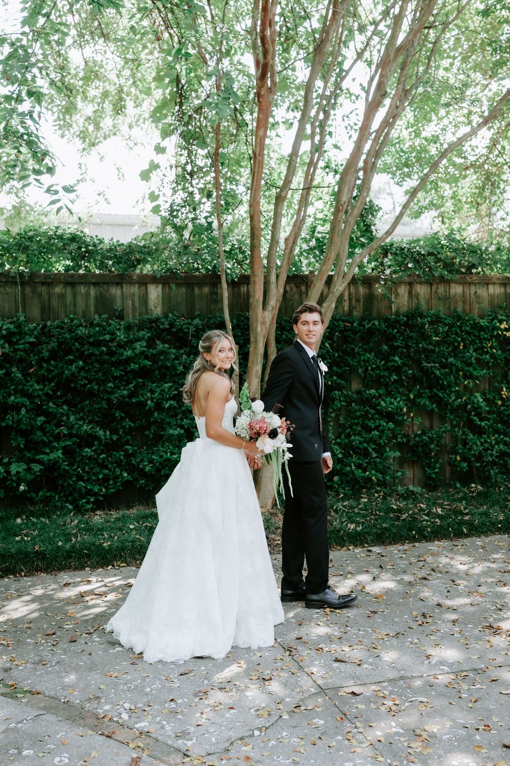 Bride in white gown and groom in black tuxedo stand back-to-back on stone patio, surrounded by lush green foliage.
