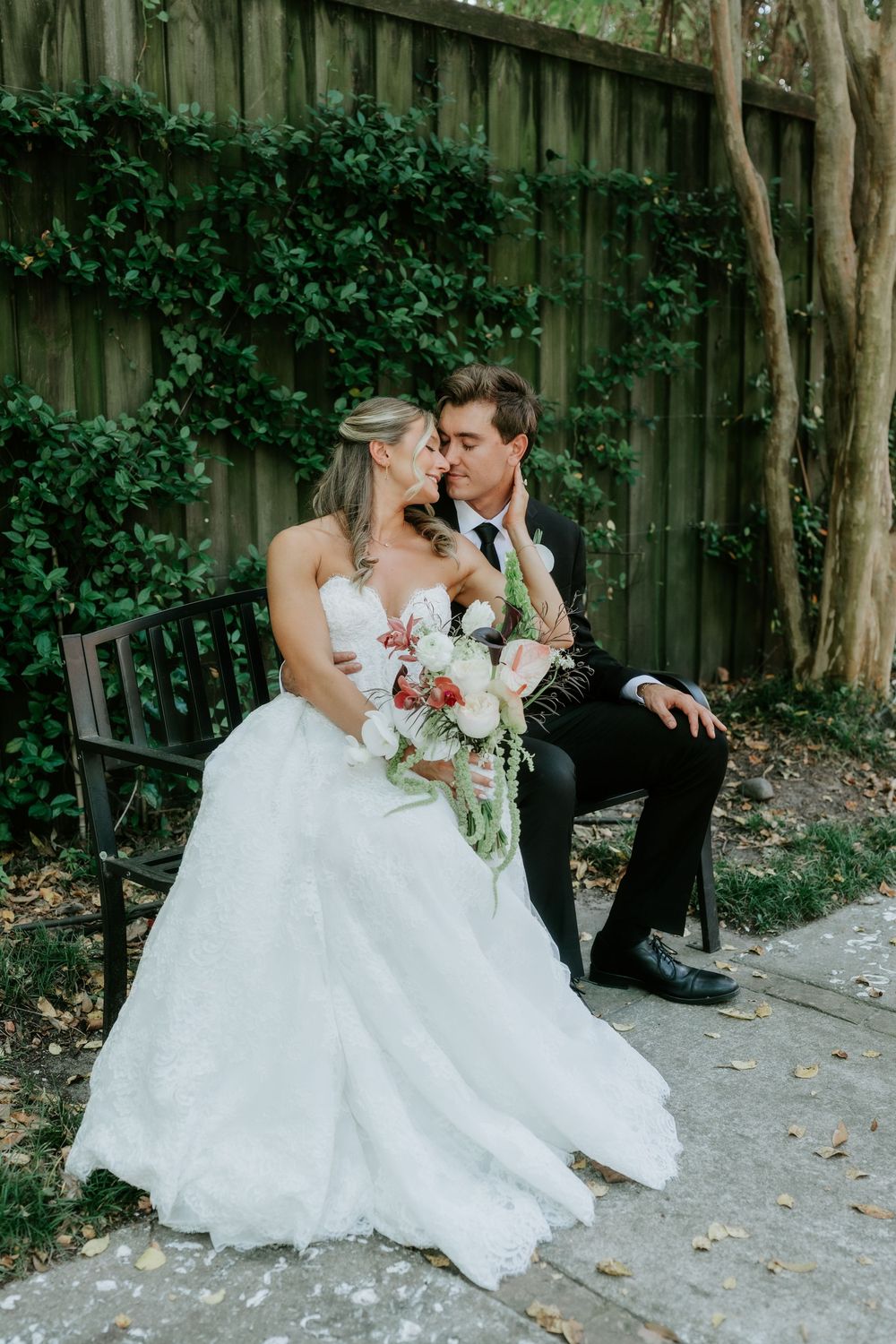 Bride and groom share a tender moment on a garden bench, surrounded by lush ivy, bride holding a pink and white bouquet.