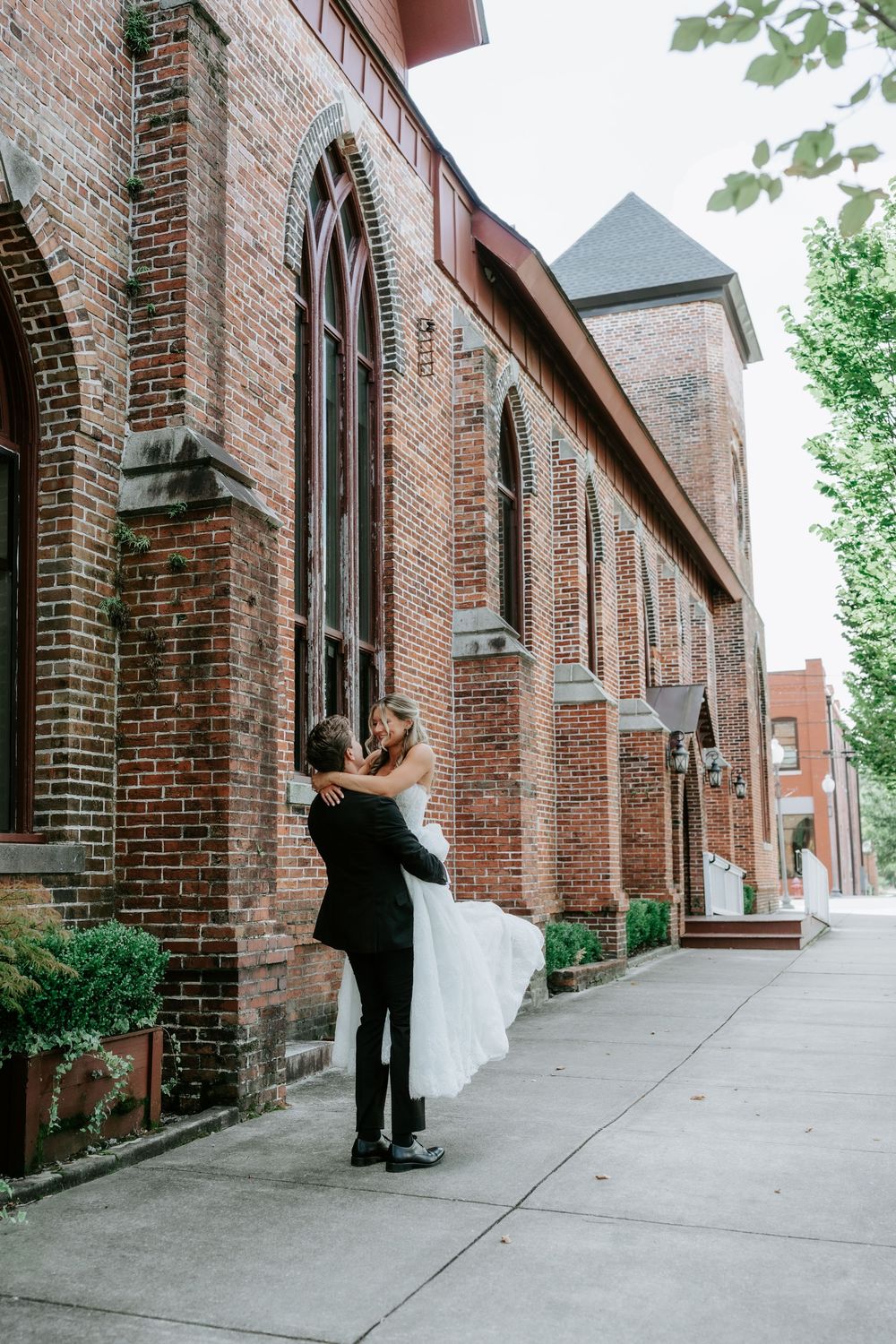Groom lifts bride in a romantic embrace outside a historic red brick church with Gothic arched windows.